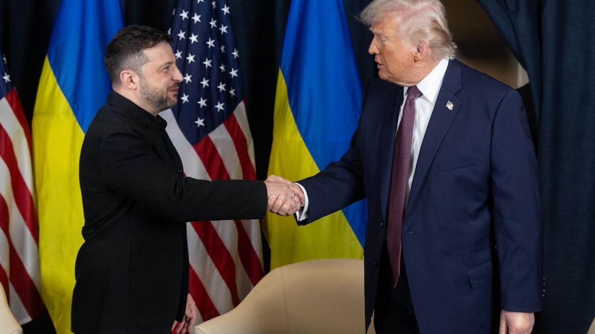 Two men in suits shake hands on a stage with American and Ukrainian flags behind them.
