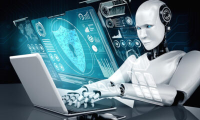 Humanoid robot typing on a laptop with glowing blue holographic security and data dashboards in front of it.