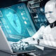 Humanoid robot typing on a laptop with glowing blue holographic security and data dashboards in front of it.