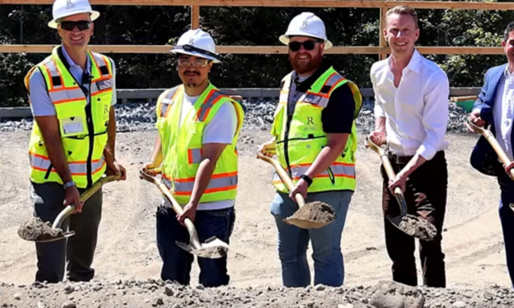 Six people in safety vests and hard hats stand with shovels on a gravel construction site.