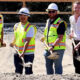 Six people in safety vests and hard hats stand with shovels on a gravel construction site.