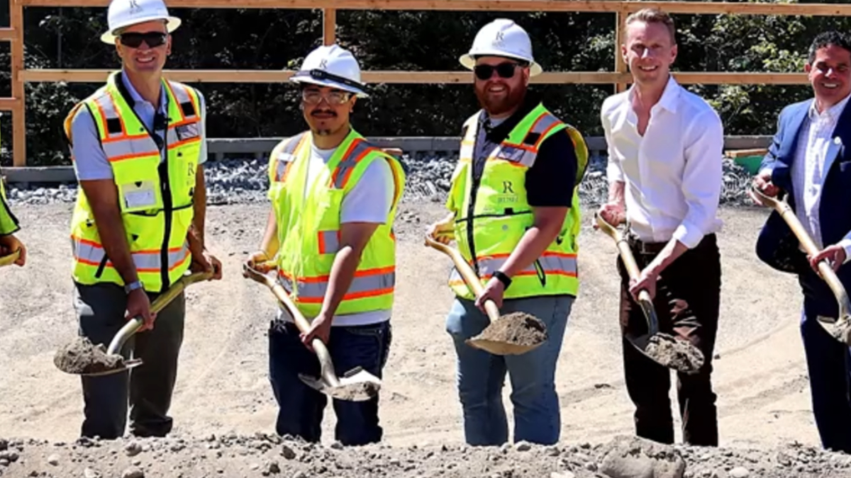 Six people in safety vests and hard hats stand with shovels on a gravel construction site.