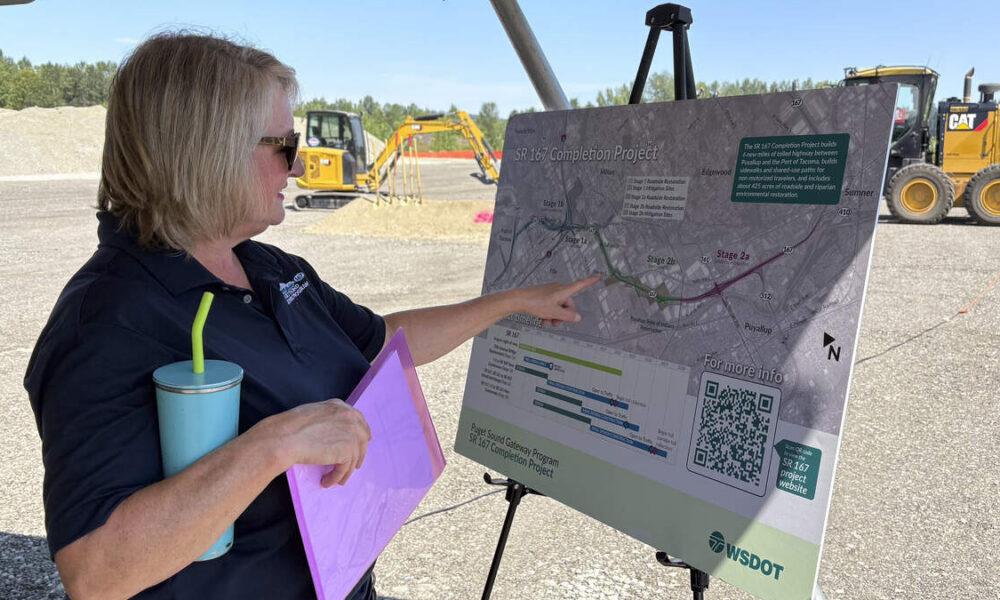 Woman in a dark polo points to a project map on a display board at an outdoor construction site, holding a purple folder and a blue tumbler with a straw.