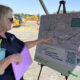 Woman in a dark polo points to a project map on a display board at an outdoor construction site, holding a purple folder and a blue tumbler with a straw.