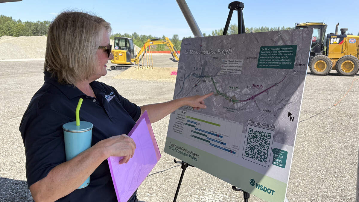 Woman in a dark polo points to a project map on a display board at an outdoor construction site, holding a purple folder and a blue tumbler with a straw.