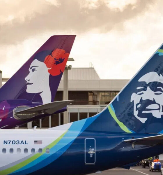 Two Alaska Airlines jets at the gate with vibrant tail art—one tail shows a woman’s profile, the other a stylized face, against a bright airport backdrop.
