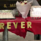 Table covered in red cloth with gold glitter letters spelling 'FOREVER 13', a wedding-style cake under a plastic cover, bouquet, decorative framed photo, and a lit decorative '2' at a celebration setup.