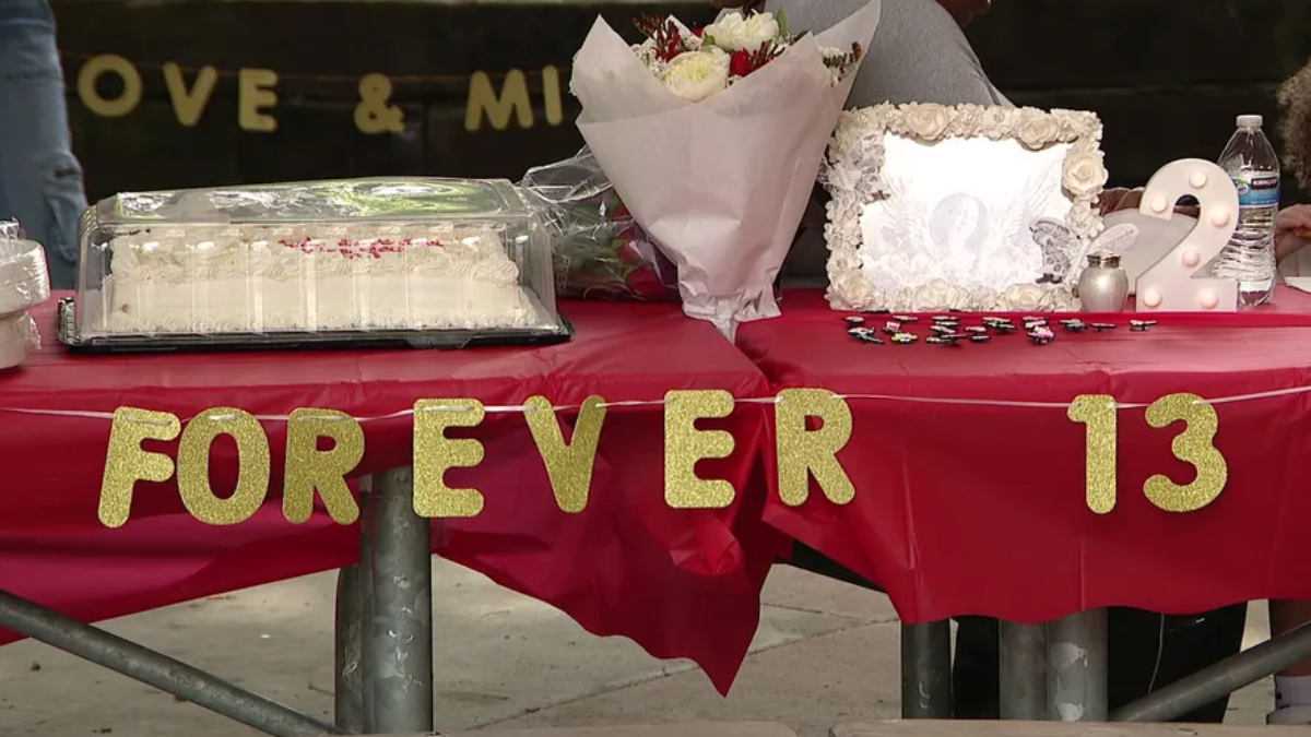 Table covered in red cloth with gold glitter letters spelling 'FOREVER 13', a wedding-style cake under a plastic cover, bouquet, decorative framed photo, and a lit decorative '2' at a celebration setup.