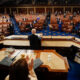 Congressional chamber: a leader stands at the central podium addressing seated lawmakers wearing masks in a semicircular chamber.