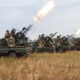 Line of military pickup trucks with soldiers manning artillery, firing in an open field with smoke plumes.