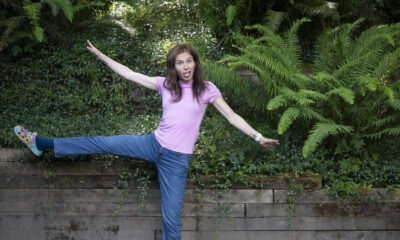 Woman in a pink shirt and jeans balances on one leg with arms outstretched in a lush garden setting.