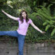 Woman in a pink shirt and jeans balances on one leg with arms outstretched in a lush garden setting.