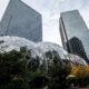Geodesic glass domes in a modern city plaza surrounded by tall glass skyscrapers and trees.