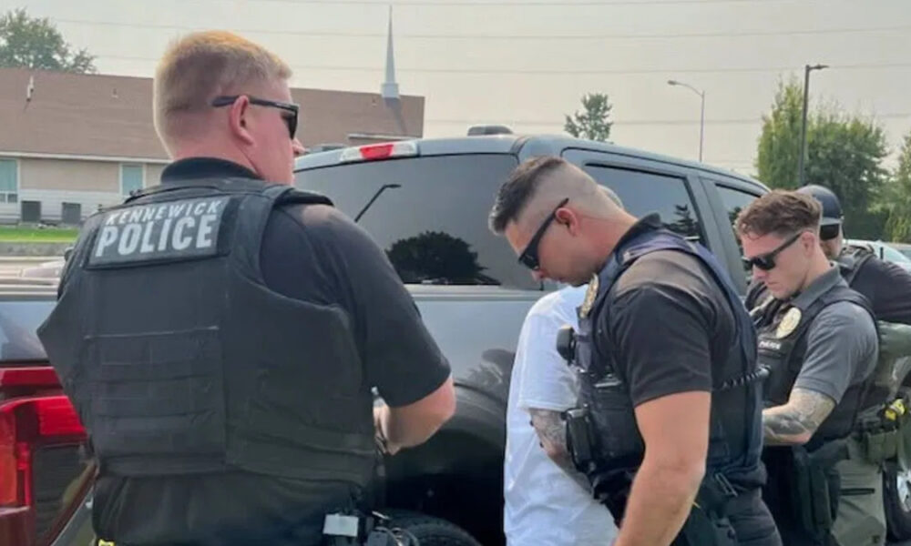 Three officers in tactical vests detain a person next to a dark SUV in a parking lot. Hidden church steeple in the background suggests a suburban area.