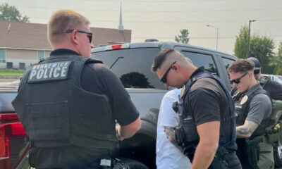 Three officers in tactical vests detain a person next to a dark SUV in a parking lot. Hidden church steeple in the background suggests a suburban area.