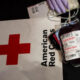 Blood donation setup: labeled blood bag with tubing on a surface beside the American Red Cross logo. Gloved hands hold the tubing nearby.