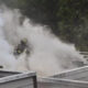 Firefighter in turnout gear on a rooftop amid thick white smoke from a fire.”, “Firefighter battles rooftop blaze, dense steam obscures view near railing.”, “Emergency responder in full gear on roof as smoke pours around.”, “Firefighter using water on a roof with heavy steam and smoke.”, “Firefighter on a flat roof with nearby trees, smoke billowing.”