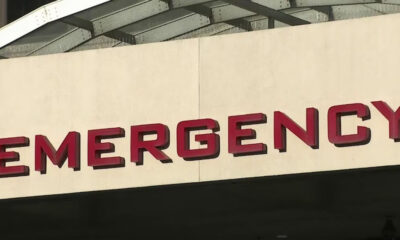 Partial view of a beige wall with red letters spelling EMERGENCY above an entrance, with a curved white canopy overhead (emergency entrance sign).