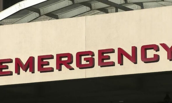 Partial view of a beige wall with red letters spelling EMERGENCY above an entrance, with a curved white canopy overhead (emergency entrance sign).