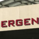 Partial view of a beige wall with red letters spelling EMERGENCY above an entrance, with a curved white canopy overhead (emergency entrance sign).