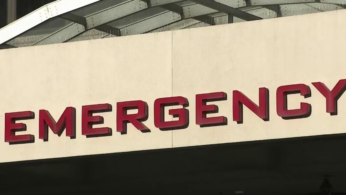 Partial view of a beige wall with red letters spelling EMERGENCY above an entrance, with a curved white canopy overhead (emergency entrance sign).