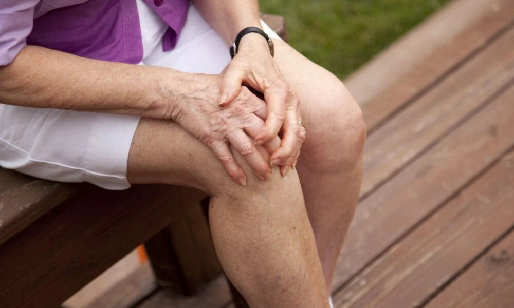 An older person sits on a wooden bench, hands clasped on their knee.