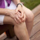 An older person sits on a wooden bench, hands clasped on their knee.