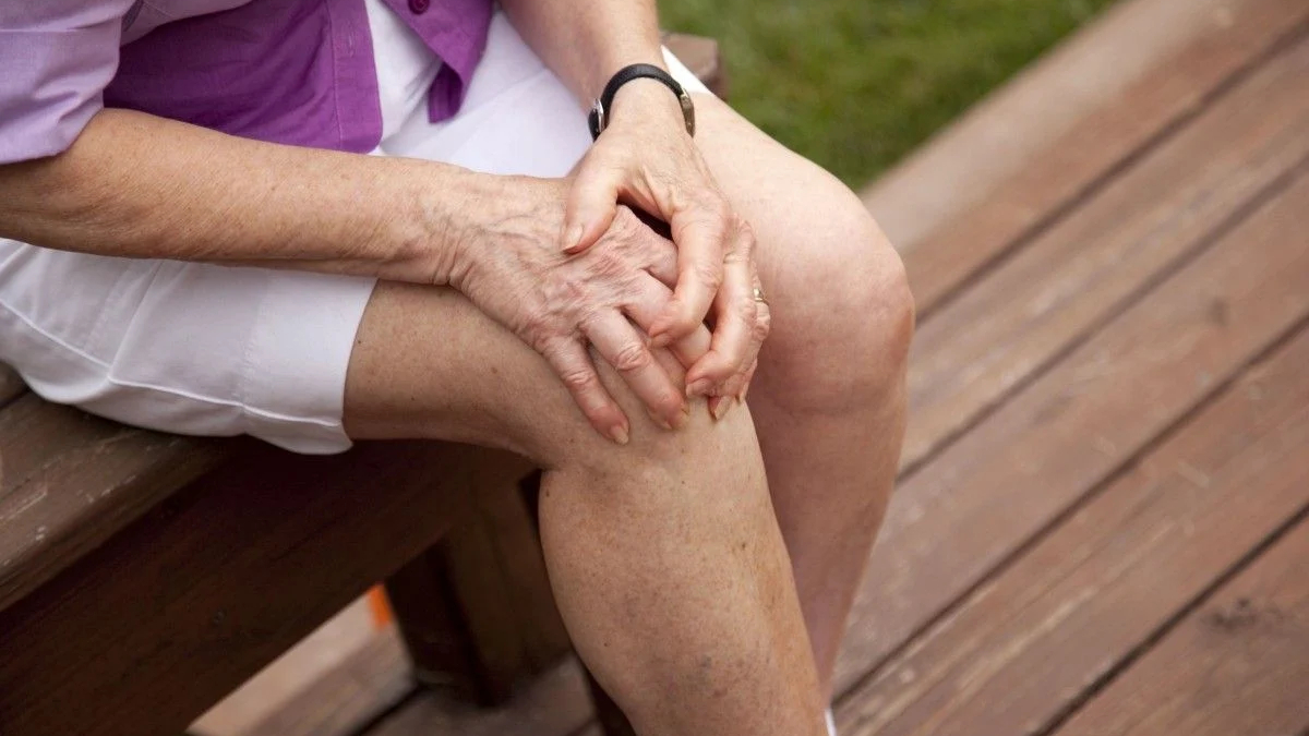 An older person sits on a wooden bench, hands clasped on their knee.