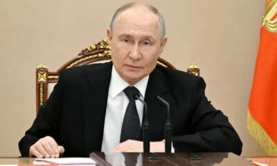 A man in a black suit and white shirt sits at a polished desk with two gooseneck microphones in front, looking toward the camera.
