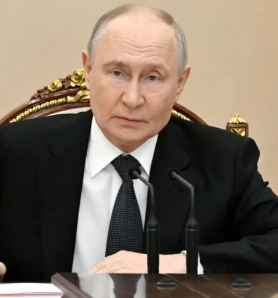 A man in a black suit and white shirt sits at a polished desk with two gooseneck microphones in front, looking toward the camera.