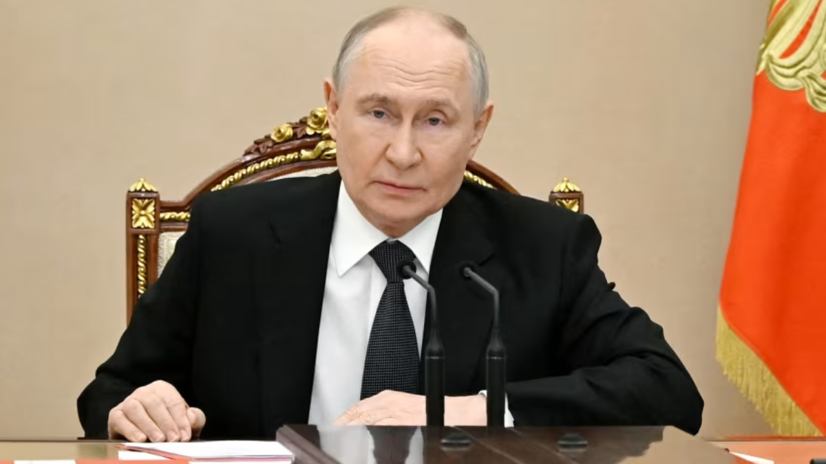 A man in a black suit and white shirt sits at a polished desk with two gooseneck microphones in front, looking toward the camera.