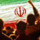 Crowd of people at a concert or rally with a large Iranian flag graphic spanning the scene, showing green, white, red stripes and the emblem in the center.