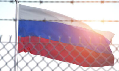 Russian flag waving behind a chain-link fence with barbed wire, sun flare in the background