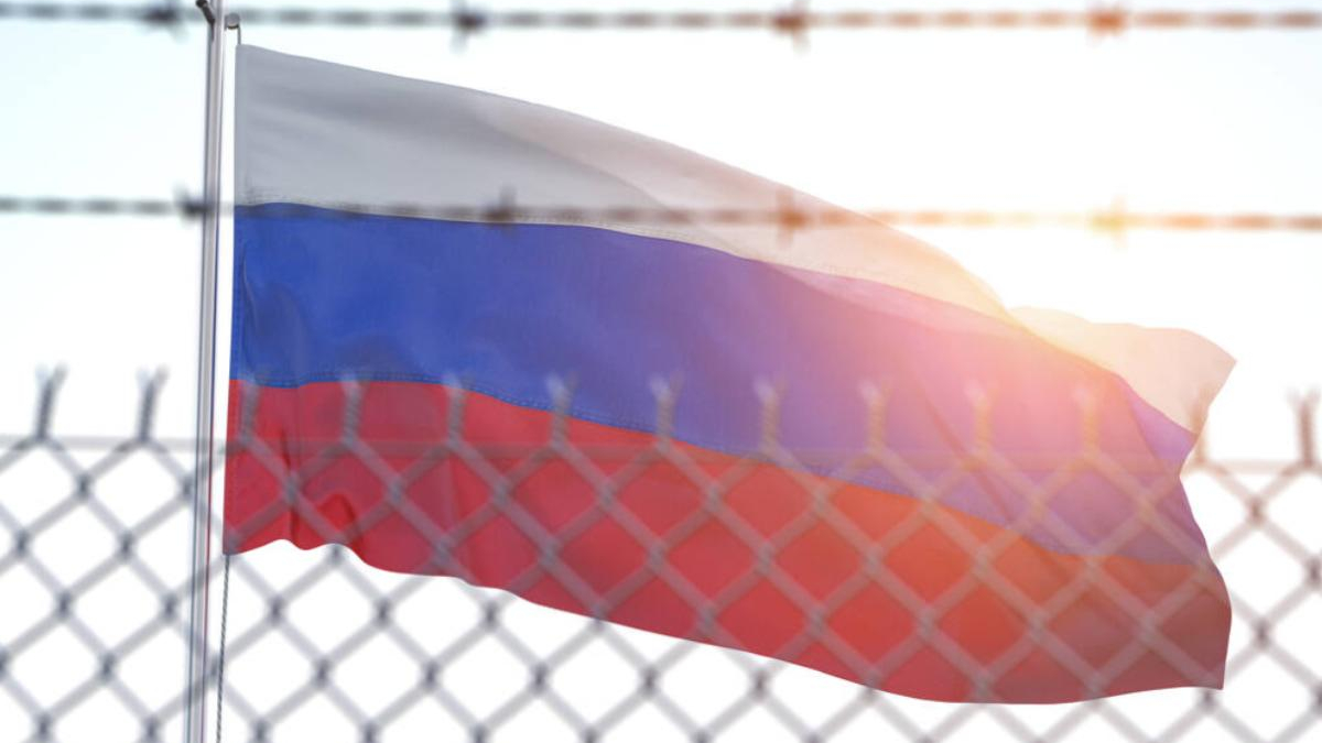 Russian flag waving behind a chain-link fence with barbed wire, sun flare in the background