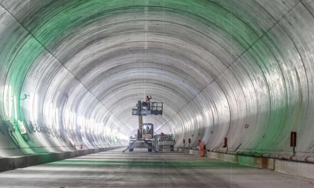 Inside a large cylindrical tunnel under construction, a scissor lift with a worker elevated near equipment.