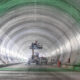 Inside a large cylindrical tunnel under construction, a scissor lift with a worker elevated near equipment.