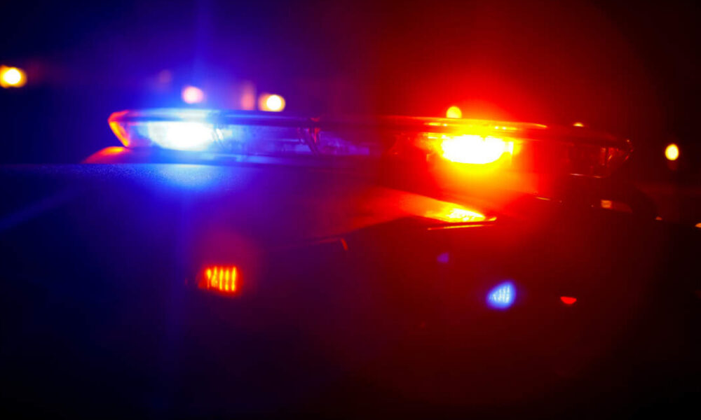 Police car with red and blue emergency lights flashing at night, horizon darkened with blurred city lights in the background