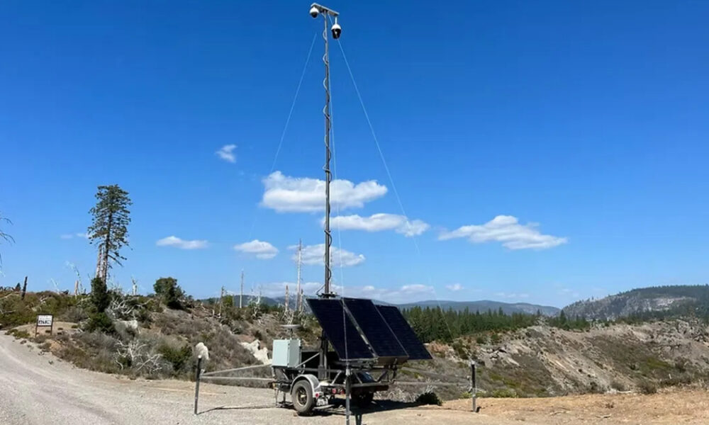 Solar-powered communication tower on a trailer with two cameras at the top, set in a dry, hilly landscape.
