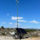 Solar-powered communication tower on a trailer with two cameras at the top, set in a dry, hilly landscape.