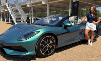 Teal convertible sports car on display with a woman in white shorts posing beside it outdoors.