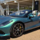 Teal convertible sports car on display with a woman in white shorts posing beside it outdoors.