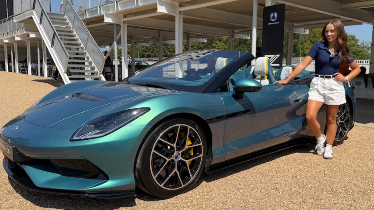 Teal convertible sports car on display with a woman in white shorts posing beside it outdoors.