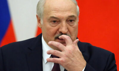 Older man in a dark suit and burgundy tie, touching his lips with his fingers against a red background with a flag edge visible.