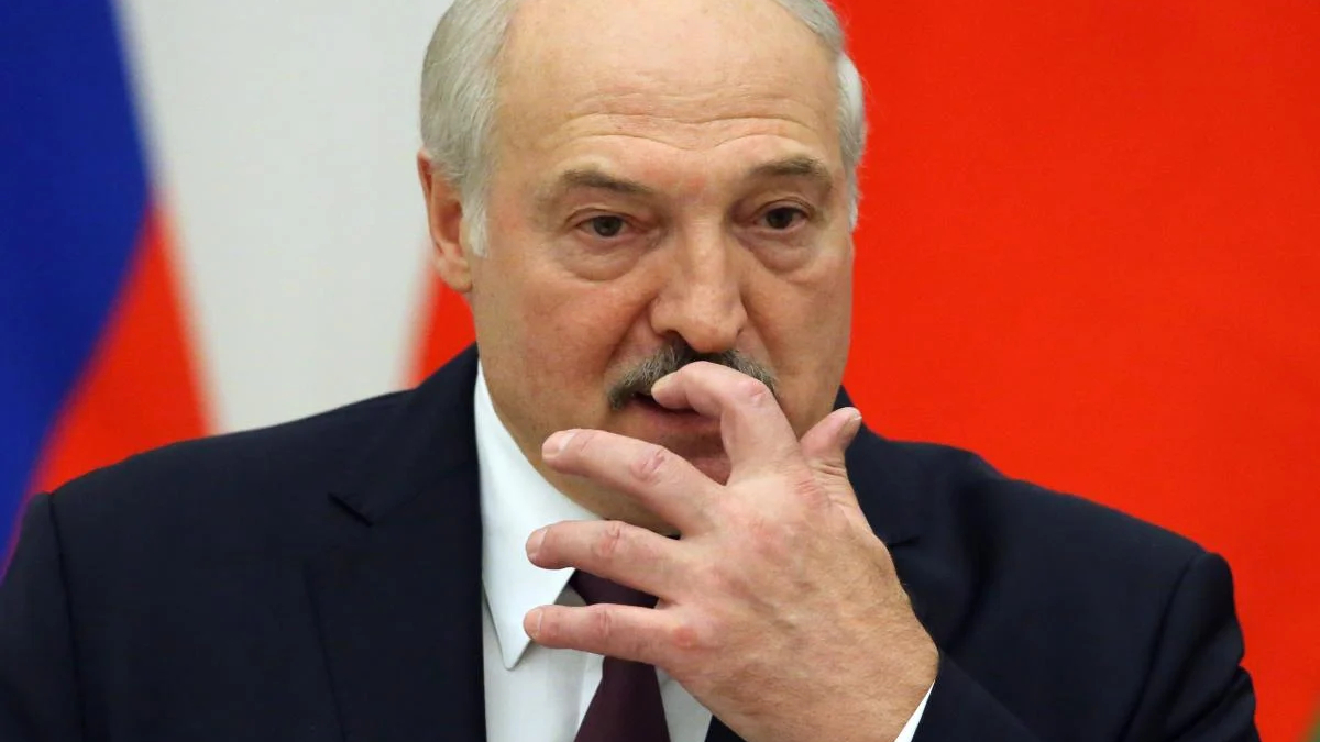 Older man in a dark suit and burgundy tie, touching his lips with his fingers against a red background with a flag edge visible.