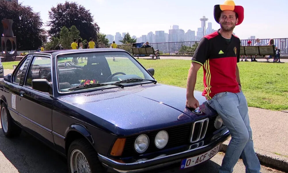 Man in a red and black jersey and a yellow-red cowboy hat leans on a dark blue vintage BMW in a park, with a city skyline in the background.