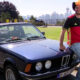 Man in a red and black jersey and a yellow-red cowboy hat leans on a dark blue vintage BMW in a park, with a city skyline in the background.