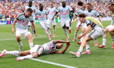 Soccer players in pastel uniforms celebrate on a green pitch; one player lies on the grass reaching out as teammates cheer.
