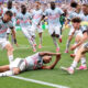 Soccer players in pastel uniforms celebrate on a green pitch; one player lies on the grass reaching out as teammates cheer.