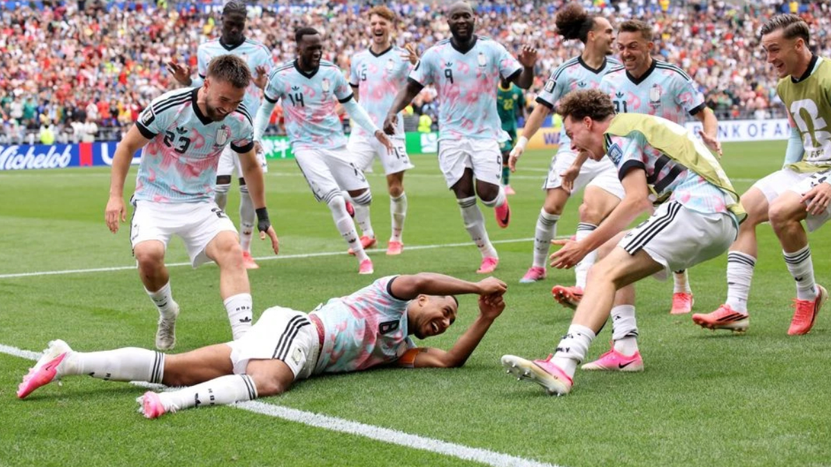 Soccer players in pastel uniforms celebrate on a green pitch; one player lies on the grass reaching out as teammates cheer.
