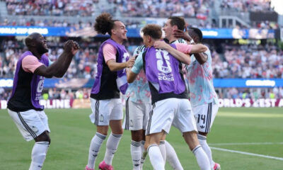Soccer players in purple bibs celebrate together on the field as fans watch from the stands behind them.